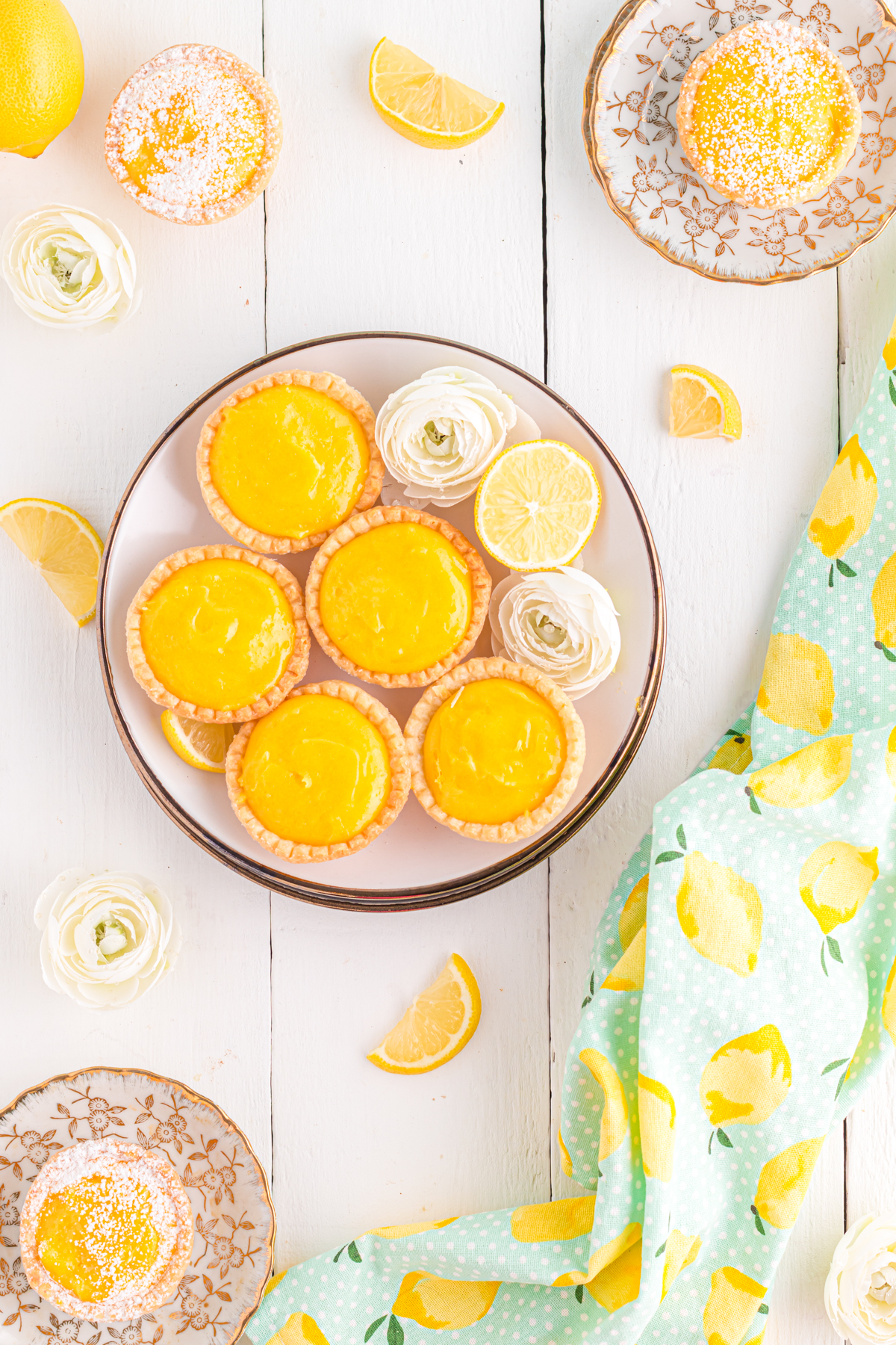 Mini lemon tarts on table with fresh lemon slices and flowers.