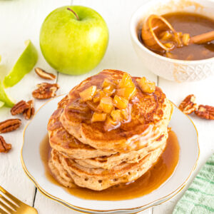Caramel Apple pancakes stacked on a plate with pecans and caramel sauce for drizzling.