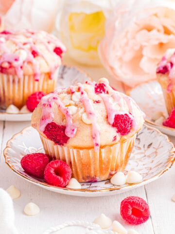 Raspberry white chocolate muffins on floral plates surrounded by raspberries and white chocolate chips.